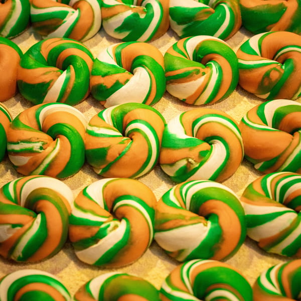 Rows of colorful green, white, and orange swirled bagels on a light-colored surface.