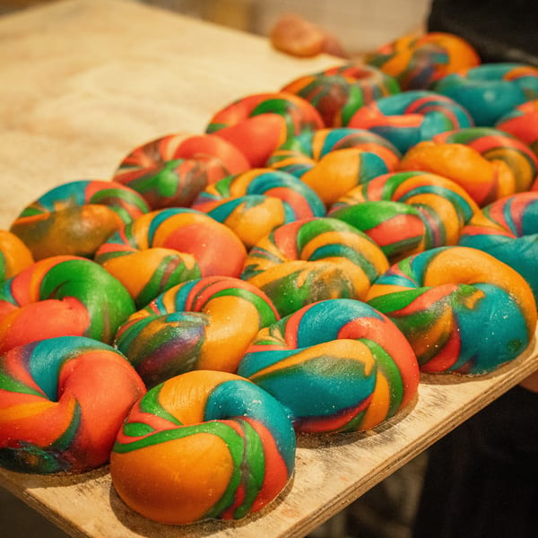 Many colorful rainbow bagels, swirled with red, orange, green, and blue, on a wooden tray.